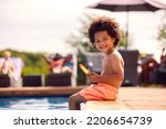 Family On Summer Holiday With Boy Eating Ice Lolly Splashing At Edge Of Swimming Pool