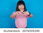 young asian woman wearing pink t-shirt against blue background has rejection angry expression crossing fingers doing negative sign.