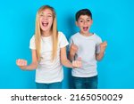 Small photo of Portrait of two kids boy and girl standing over blue background looks with excitement at camera, keeps hands raised over head, notices something unexpected reacts on sudden news.