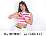 Young beautiful brunette woman wearing striped crop top over white wall smiling in love showing heart symbol and shape with hands. Romantic concept.