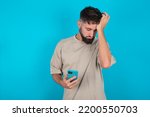 Small photo of Upset depressed bearded caucasian man wearing casual T-shirt over blue background makes face palm as forgot about something important holds mobile phone expresses sorrow and regret blames