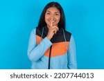 Small photo of Smiling Young latin woman wearing sport clothes over blue background makes shush gesture, holds fore finger over lips hides secret. Be mute, please.