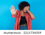 Small photo of brunette arab woman wearing pink shirt over blue background covers eyes with palm and doing stop gesture, tries to hide. Don't look at me, I don't want to see, feels ashamed or scared.