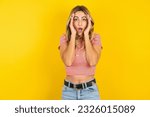 Young beautiful blonde woman wearing striped t-shirt over yellow studio background with scared expression, keeps hands on head, jaw dropped, has terrific expression. Omg concept