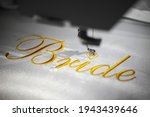 Machine embroider the inscription bride on a white satin robe.
Inscription bride with gold thread. Wedding day preparation. Elegant bridal lingerie. White accessories for the bride. Glamorous style.