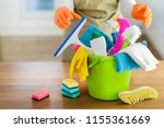 Small photo of Cleaning items on wooden table in modern kitchen. Woman house work or charwoman blured in background. Bucket, brush, washcloth, spray.