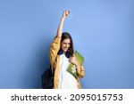 Small photo of Happy student girl says yes, passed an important test, raises hand with fist in excitement, smiles sincerely, holding books, wearing yellow shirt, white t-shirt, black bag and headphones over neck