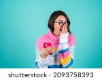 Small photo of A young woman in a bright multicolored sweater on a blue background with glasses, holds the phone, laughs cheerfully, read a joke, anecdote, covers her mouth with her hand, laughs nonstop