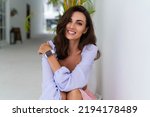 Small photo of Stylish young woman with voluminous hair in a trendy long sleeve crop top and a pink skirt posing against a white wall and a tropical bush, wearing a smart watch