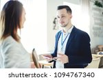 Small photo of Confident young businessman with tablet computer in his hands and accreditation badge around his neck talking with unrecognizable female colleague at business event