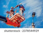 Small photo of Two electricians from cradle of aerial platform or crane are repairing street lighting lamp. Professional electricians wearing helmets, overalls and insurance work at heights. View of workers from