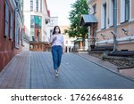 Caucasian young woman in protective mask on her face walks alone along the street, listens to music. Concept: solo walks great outdoors in uninhabited places. Europe. City environment. Summer morning.