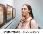 Portrait of young caucasian thoughtful woman looks at the painting. Exhibition in the art gallery. Excursion and visit to the museum.