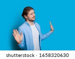 Small photo of Bearded caucasian man with long hair is gesturing the blameless sign with his hands while posing on a blue wall