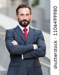 Small photo of Confident pleasant unshaken businessman standing outside office smiling and holding hands crossed. I am professional. portrait of successful businessman in suit and red tie