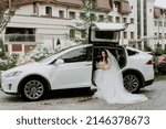 Stylish Bride with Flower Bouquet near Limousine. Pretty Young Woman Dressed White Wedding Dress and Jewelry Diadem with Veil. Girl Holding Roses Bunch and Standing near white car. Outside Photo