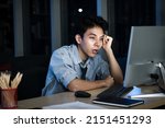 Small photo of Asian young tired staff officer man using desktop computer having overwork project overnight in office, exhausted unhappy businessman feeling sleepy after after working hard overtime at night.