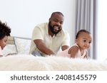 Small photo of Happy African family, little smiling cute toddle baby infant daughter girl crawling on white bed in bedroom with blurred background of her father and son brother. Kid and parent spending time together