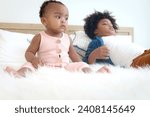 Small photo of African cute toddle baby infant girl sitting white her relaxing brother boy lying down on white bed in bedroom. Portrait of adorable little kid at home. Happy African child siblings in family.