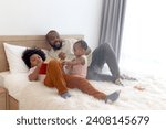 Small photo of Happy African family, little cute toddle baby infant daughter girl plays with father on white bed while son brother reads in bedroom. Dad looks after child at home, kids and parent spend time together