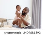 Small photo of Happy African family, father holding his little cute toddle baby infant daughter girl while sitting on white bed in bedroom. Dad looking after child at home, kid with parent spending time together.