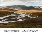 Wide view over a flat valley in the highlands of Iceland, a river winds with many river branches through the landscape shining in brown and orange tones, cloud shadows 