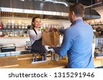Cheerful waitress wearing apron serving customer at counter in restaurant - Small business and service concept with young business owner woman giving bag with takeaway food to client