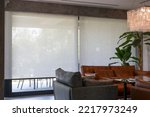 Roller blinds in the interior. Automatic solar shades large size on the window. Living room interior with sofas and palm trees. Electric sunscreen curtains for home. 