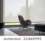Roller blinds of large sizes on the window in the interior. Automatic solar shades, fabric with linen texture. In front of a large window is a chair on a carpet. Outside is a view of the garden.