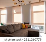 Small photo of Motorized roller blinds. A sofa with colorful pillows in the room near windows with sunscreen curtain. Automatic roller shades on full height windows in the interior. Sunny day. Selective fokus.