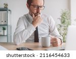 Small photo of Portrait of perturbed businessman with cup of coffee in the office looking at laptop computer screen and thinking, selective focus