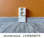 Small photo of Electric socket by the floor between plastic baseboard with a wooden texture. Newly installed wooden grey carpet flooring and baseboards in home. Close-up of plastic plinths and caramel wall.