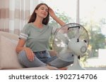 Woman enjoying air flow from fan on sofa in living room. Summer heat