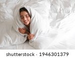 Cheerful young woman covered with warm white blanket lying in bed. Space for text