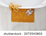 Small photo of Soft orange bath mat and slippers on floor in bathroom, top view