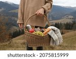 Woman holding wicker picnic basket with thermos, snacks and plaid in mountains on autumn day, closeup