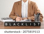 Small photo of Black cubes with word Blacklist and woman working at wooden office desk, closeup