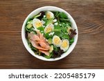 Delicious salad with boiled eggs, salmon and cheese in bowl on wooden table, top view