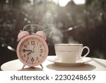 Pink alarm clock and cup with hot drink on white table in morning