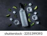 Bottle of vodka, shot glasses, lime, mint and ice on black table, flat lay