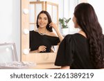 Young woman trying on elegant pearl earring near mirror indoors