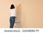 Small photo of Young woman painting wall with roller on stepladder indoors, space for text. Room renovation