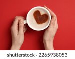 Woman holding cup of aromatic coffee with heart shaped decoration on red background, top view