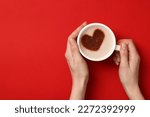Woman holding cup of aromatic coffee with heart shaped decoration on red background, top view and space for text