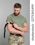 Small photo of Soldier in military uniform applying medical tourniquet on arm against light grey background