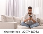 Man using smartphone on sofa near window with beautiful curtains in living room. Space for text