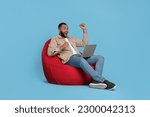 Small photo of Happy young man with laptop sitting on beanbag chair against light blue background