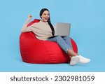 Small photo of Cheerful woman with laptop sitting on beanbag chair against light blue background