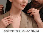 Man putting elegant necklace on beautiful woman against dark grey background, closeup