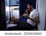 Man eating sausages near refrigerator in kitchen at night. Bad habit